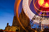 The glowing Ferris wheel spins by the Tibidabo church at twilight, highlighting the amusement parks vibrant colors.