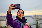 Young woman taking a selfie with skyline behind, including Parliament building and Danube River, Budapest, Hungary, Europe