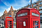 Two iconic red telephone boxes stand on Brompton Road near Harrods, framed by a bright blue sky and bustling city life.