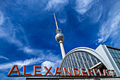 The iconic Alexanderplatz station sign stands prominently with the Fernsehturm rising in the background against a blue sky.