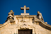 The ornate top of Santa Cecilia Church features a cross and statues, set against a clear blue sky in Ronda, Andalusia.