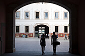 Two individuals walk through an archway into the vibrant patio area of the cultural center, surrounded by architecture.