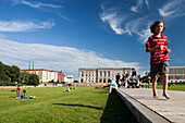 Children play on the lawn at Schlossplatz, with the distinctive Council of State Building in the background under a clear blue sky.