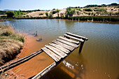 An old wooden dock extends into the tranquil waters of Lake Sumido, surrounded by the natural landscape of Las Médulas.