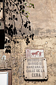 A weathered street sign identifies Placa de la Calle de la Cera in the vibrant Raval neighborhood of Barcelona under leafy branches.