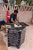 A young man grills traditional tortillas rellenas for sale on a cobblestone street in Purmamarca, Argentina.