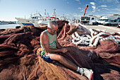 Seville, Spain, Aug 7 2008, A fisherman repairs his nets at Bonanza port in Sanlucar de Barrameda, Cadiz, while boats and vehicles surround him under a clear sky.