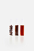 Three small glass bottles against a white background, filled with different types of coffee; the first contains whole coffee beans, the second ground coffee and the third liquid coffee extract