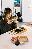 Young woman enjoying food and wine at home and taking photos with her mobile phone