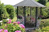 Pavillon mit Eisenstühlen, Rosen (Rosa) und Blumenkästen vor Pfingstrosen (Paeonia) im Sommergarten