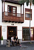 Spain, Canary Islands, Palma Island, couple sitting on a bench in front of the wooden balcony of a Spanish colonial house in the historic center of Santa Cruz de la Palma