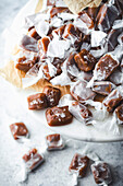 A close-up of a large stack of homemade caramel sweets on a marble surface. The candies are wrapped in white parchment paper and sprinkled with sea salt. They are arranged on a piece of brown parchment paper standing on a white marble cake stand. More caramels can be seen in the blurred background, hinting at a delicious treat and emphasising the simplicity and beauty of homemade sweets