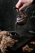 Chinese tea ceremony. A man's hand pours tea from a traditional handmade black ceramic pot with a wooden handle into a cup on a dark tea table. Autumnal atmosphere. Dried flowers bloom nearby