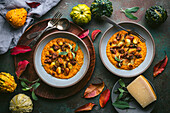 A wide shot of two bowls of crispy gnocchi in pumpkin sauce with sage and sausage arranged on a dark green surface. The bowls are surrounded by fall decor, including gourds, leaves, and a sprig of sage. A wooden tray holds silverware, and on the other side is a small plate with a piece of Parmesan cheese. The image is a beautiful and appetizing representation of fall flavors.