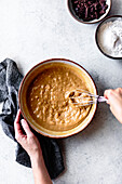 A person beats a creamy batter in a bowl, surrounded by chocolate chips and flour, with a kitchen towel nearby