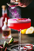 An action shot of bright pink berry powder being sprinkled on sophisticated alcoholic beverage, featuring a textured stemmed coupe cocktail glass with colorful red liquor with a foamy top