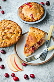 A close-up of 2 freshly baked mini apple and cranberry cakes. One piece is on a small plate. Apple slices and cranberries are scattered on the plate. The pie is golden brown and the filling is visible through the crust