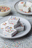 Side view of a stack of square white marshmallows sprinkled with colourful rainbow sprinkles on a light grey plate. A soft focus bowl with sprinkles and more marshmallows in the background. A cheerful and colourful shot, light and bright