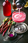 Action shot of pink rhubarb cocktail being poured from shaker into beautiful half-full coupe glass with fresh rhubarb and red syrup in background