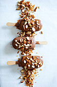 Three almond ice-cream sticks covered in milk chocolate, sprinkled with roasted almonds on a white marble base