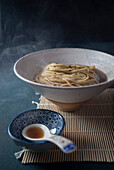 A steaming bowl of ramen noodles in broth,garnished with black sesame seeds,along with a side dish in a blue patterned bowl,on a bamboo mat,against a dark background