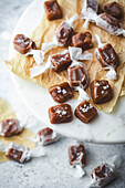 A close-up of homemade caramel sweets on a marble surface. The sweets are wrapped in white parchment paper and sprinkled with sea salt. Arranged on a piece of brown parchment paper standing on a white marble cake stand. In the blurred background are more caramels, hinting at a delicious treat and highlighting the simplicity and beauty of homemade sweets