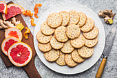 A white plate with homemade ginger and grapefruit biscuits on a grey background. Slices of fresh pink grapefruit and ginger root lie on a wooden chopping board. Citrus peel is scattered across the board. A knife with a wooden handle lies on the worktop next to the plate. This picture conveys a feeling of home-made cosiness and the delicious taste of citrus fruits and ginger
