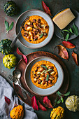 A wide shot of two bowls of crispy gnocchi in pumpkin sauce with sage and sausage arranged on a dark green surface. The bowls are surrounded by fall decor, including gourds, leaves, and a sprig of sage. A wooden tray holds silverware, and on the other side is a small plate with a piece of Parmesan cheese. The image is a beautiful and appetizing representation of fall flavors.