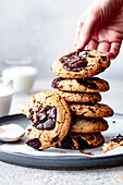 A hand reaches for a freshly baked chocolate chip cookie from a stack on a gray plate, with a glass of milk in the background.