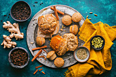 A rustic, teal-colored background with a variety of chai spices and freshly baked muffins, some whole and some broken apart, revealing their fluffy interior. Around the platter are cinnamon sticks, fresh ginger, and small bowls filled with various spices, including black peppercorns, cloves, fennel seeds, and cardamom pods, adding a touch of natural color and texture. A saffron yellow linen napkin adds a pop of warmth and complements the overall rustic aesthetic. The image captures a simple, yet inviting scene, suggesting the warmth of home baking and the comforting aroma of spices.