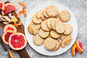 A white plate with homemade ginger and grapefruit biscuits, some of which are broken, on a custom grey background. Slices of fresh pink grapefruit and ginger root lie on a wooden chopping board. Citrus peels are scattered across the board. A knife with a wooden handle lies on the worktop next to the plate. This image conveys a feeling of home-made cosiness and the delicious taste of citrus fruit and ginger