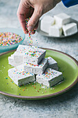 A hand sprinkles colourful sprinkles on a pile of square white marshmallows stacked on a green ceramic plate. A soft-focus bowl with sprinkles and more marshmallows in the background. A cheerful and colourful, bright shot