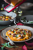 Side view of a plate of gnocchi and sausage with sage and parmesan cheese on a pumpkin sauce. One hand holds a red grater with which the parmesan is sprinkled over the dish. A rustic, autumnal table setting can be seen in the background