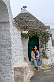 Woman tourist sit on the front of one Trullo traditional house with conic shape ceiling, UNESCO World Heritage Site, Alberobello, Apulia, Italy, Europe