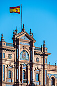 Close up of the intricate architectural details of Plaza de España in Seville, Spain, featuring traditional Spanish design elements and a vibrant flag against a clear blue sky.