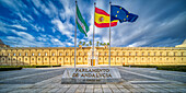 The image captures the iconic Hospital de las Cinco Llagas, now the Parliament of Andalusia, in Seville. Three flags wave prominently under a vivid sky, showcasing cultural and historical significance.