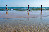 Three individuals walking along the shoreline at Playa de Cuesta Maneli in Huelva, Andalucia. The serene beach scenery captures movement and reflection under a clear sky.