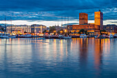 Stunning evening view of Oslo's City Hall and Aker Brygge from Akershus docks, with illuminated buildings reflecting on calm waters under a dramatic sky.