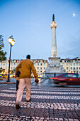 Ein Fußgänger überquert am Abend den Zebrastreifen am Rossio-Platz in Lissabon,während Autos vorbeifahren und ein historisches Denkmal in der Nähe steht.