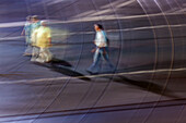 Artistic long-exposure photograph capturing the dynamic motion of people walking in the city at night. The image presents a blur effect conveying movement and energy.