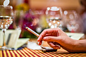 Close up of a woman's hand holding a cigarette at a restaurant, capturing a sophisticated, relaxed atmosphere with soft lighting and wine glasses in the background.