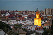 Aerial view of the illuminated mudejar bell tower of San Marcos Church in Seville, Spain, surrounded by urban architecture and vibrant cityscape during dusk.