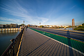 Seville, Spain, Jan 28 2021, Wide angle view of Triana Bridge over Guadalquivir River in Seville, Spain. Torre Sevilla in the background under a clear blue sky. Shadow patterns create a striking visual on the walkway.