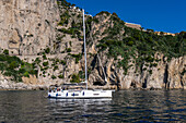 A sailboat anchored in the waters of the Tyrrhenian Sea off the coast of the island of Capri, Italy. Hotels in the town of Capri are visible at the top with the switchbacks of Via Krupp winding down the cliff.