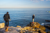 Two men enjoying fishing on a rocky shoreline overlooking the calm Mediterranean sea.