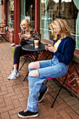 Zwei Teenager genießen die Zeit miteinander und sitzen an einem Kaffeetisch in einem Straßencafé und verbringen Zeit mit ihren Smartphones; St. Albert, Alberta, Kanada.