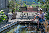 Schwimmteich und Sitzplatz auf sommerlicher Terrasse mit Olivenbaum und Hortensie im Topf 