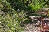 Sitzplatz mit Steinmännchen vor Beet mit Schnittlauch, Chinaschilf, Lampenputzergras und Farnblätter im sommerlichen Garten 