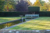 Herbstgarten mit weißer Metallbank und Gartenamphore im Teich und Hecken Formschnitt