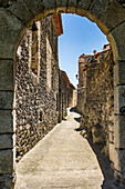Stone archway leading to a narrow historical street in Mosset, France under blue sky.